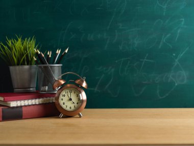 Education concept, study table with clock, stationery, plant pot and copy space on wooden table with chalkboard wall background