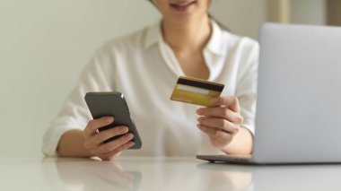 Akıllı telefondan alışveriş yapan ve çalışma yerinde otururken kredi kartı tutan bir kadının görüntüsü.