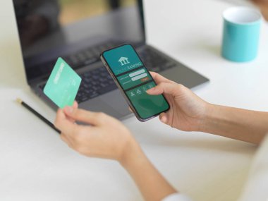 Akıllı telefonda internet bankacılığı uygulaması kullanan ve çalışma alanında kredi kartı tutan kadın elinin görüntüsünü kapat