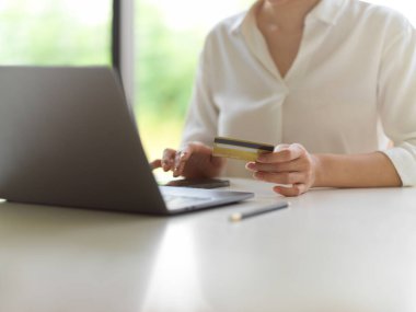 Kredi kartı tutan ve bilgisayar bilgisayarıyla beyaz masadaki akıllı telefon ekranına dokunan bir kadın eli görüntüsü. 