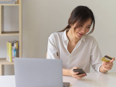 Ofis odasında online alışveriş yaparken akıllı telefon kullanan ve kredi kartı tutan bir kadının portresi