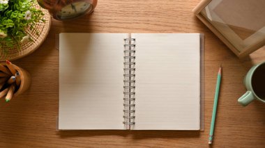 Boş defter, kalem, bardak ve süslemelerle dolu ahşap çalışma alanının üst görüntüsü.