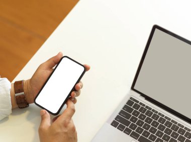 Erkek ellerinin akıllı telefonu tutarken üst görüntüsü ekranı taklit ediyor elleri beyaz masada dizüstü bilgisayarlı, patikayı kesiyor.