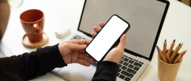 Siyah ceketli kadının elinde akıllı telefon olan görüntüsü, dizüstü bilgisayarla beyaz masada çalışırken yolu kesmeyi de içeriyor