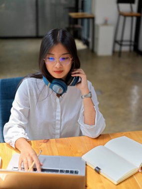 Kulaklıklı genç bir kadının portresi kütüphanede boş bir defterle ahşap masada dizüstü bilgisayar kullanıyor. 