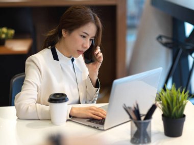 Beyaz takım elbiseli bir iş kadınının portresi telefonda konuşuyor ve ofis odasında dizüstü bilgisayarla çalışıyor.