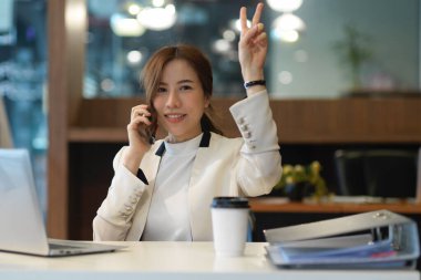 Neşeli iş kadınının yarım boy portresi telefonda konuşuyor, gülümsüyor ve iki bulucuyu kameraya gösteriyor.