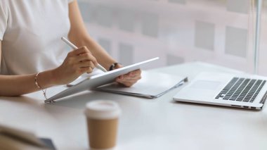 Kütüphanedeki beyaz masada ödev yapmak için dijital tablet kullanan kadın öğrenci elini kapat