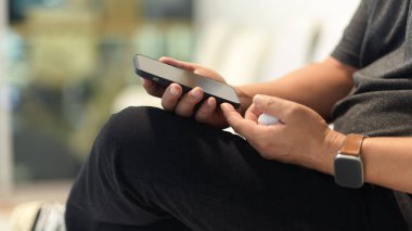 Arkaplanda bulanık otururken elinde akıllı telefon ve kablosuz kulaklık tutan bir adamın yan görüntüsü