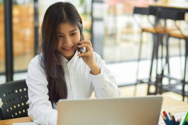 Ofiste dizüstü bilgisayarla çalışan genç bir kadın telefonda konuşuyor.