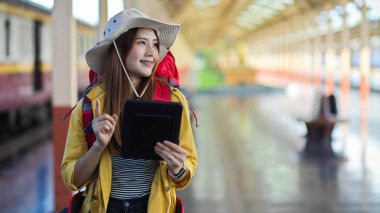 Tren istasyonunda varış yerini ararken gülümseyen genç bayan sırt çantasının yarım boy portresi.