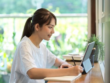 Kafede kırtasiye malzemesi ve dijital tabletle ev işi yapan kız öğrencinin yan görünümü