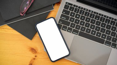 Top view of smartphone with mock-up screen on laptop on wooden workspace with notebooks