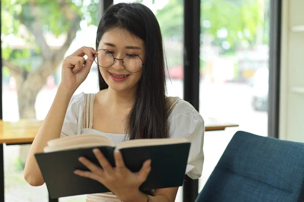 図書館に座っている間に眼鏡をかけて読書をする女性学生の笑顔の肖像 — ストック写真