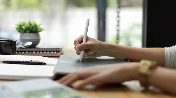 Side view of female hands using digital tablet with stylus pen on the table with stationery