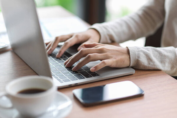 Close up of female fingers typing on laptop keyboard with smartphone and coffee on the wooden desk