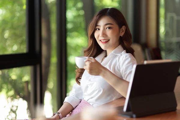 身着正装的女商务人士在笔记本电脑前喝咖啡 一边喝着好咖啡 一边欣赏着户外绿意盎然的景色 — 图库照片