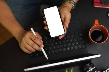 Birlikte çalışma alanındaki iş verilerini analiz etmek için akıllı telefon ve tablet kullanan serbest çalışan erkek, boş ekrandaki aygıt, modern siyah çalışma alanı