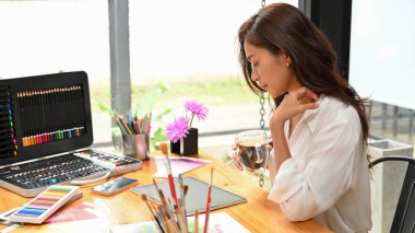 Kadın grafik tasarımcısı dijital tablet, yaratıcı ofis ve boya malzemeleriyle ilgili çalışmalarına bakarken kahve içiyor.