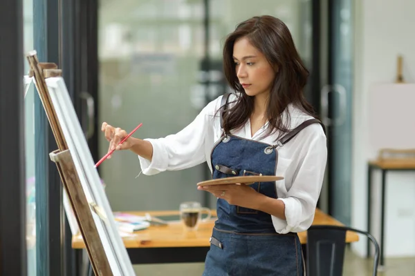 Creative hipster woman painter paints on canvas with acrylic colour, art palette in hand at workshop art studio