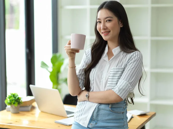 成功的年轻女商人穿着休闲时髦的服装 在现代办公室的背景下拿着咖啡杯和微笑拍照 — 图库照片