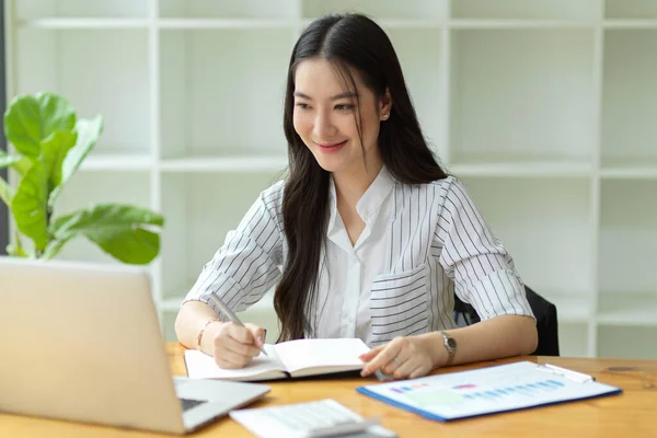 在办公室工作的年轻亚洲女商人在笔记本上写着商业计划 看了看笔记本电脑屏幕 — 图库照片