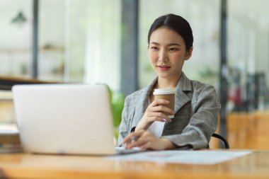 Ofiste çalışan başarılı iş kadını, dizüstü bilgisayar ekranına bakıyor, kahve fincanı tutarken iş stratejisini düşünüyor ve analiz ediyor, çalışanları rahatlatıyor.