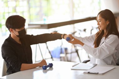 Çekici kadın terapist, erkek hastaya sağlık terapi merkezinde fizik tedavi yapması için yardım ediyor. Erkek hasta koluna bakmak için küçük dambıllar tutuyor..