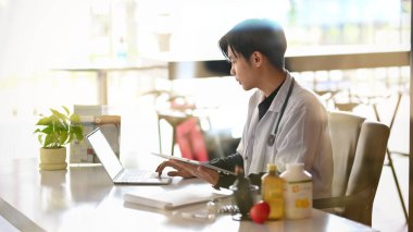 Masasındaki dizüstü bilgisayarda çalışan genç uzman doktorun yan görüntüsü. Doktorun masası.