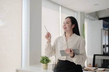 Kadın ofis çalışanı çalışma masasının önünde dururken tabletten bakıp gülümsüyor. Sanatçı ya da tasarımcı, modern şirket, özgürce düşünüyor..