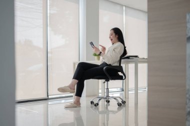 Kadın ofis çalışanı ya da iş kadını boş bir ofiste döner sandalyede otururken telefona gülüp işaret ediyor. Modern şirket bekliyor ya da dinleniyor..