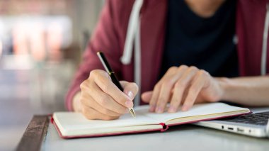 Masada dizüstü bilgisayarla otururken elinde tükenmez kalemle kırmızı bir deftere yazı yaz. Çalış ya da dışarıda çalış.