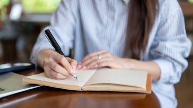 Kafedeki ahşap masada otururken not defterine dolma kalemle yazan mavi çizgili tişörtlü bir kadının fotoğrafını çek. Dışarıda çalış ya da çalış.