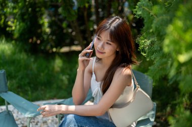 Gecelikli ve kot pantolonlu Asyalı bir kadın telefonda konuşuyor ve yeşil bahçede ya da parkta otururken ileriye bakıyor. Dinleniyorum, dışarıda dinleniyorum..