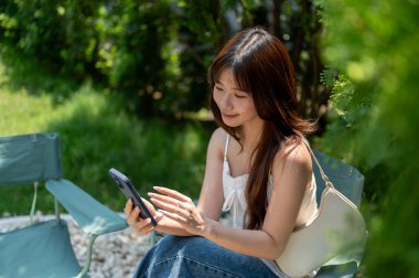 Gecelikli Asyalı bir kadın ve kot pantolon giymiş yeşil bahçede ya da parkta koltuğunda otururken telefonuna bakıp gülümsüyor. Dinleniyorum, dışarıda dinleniyorum..