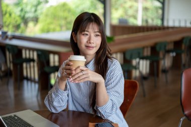 Mavi gömlekli gülümseyen Asyalı kadın kafedeki tahta masada dizüstü bilgisayarla otururken iki eliyle kahve fincanı tutuyordu. Dışarıda çalış ya da çalış.