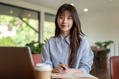 Mavi çizgili tişörtlü Asyalı kadın elinde dolmakalemle kafede dizüstü bilgisayarla otururken kameraya mutlu bir şekilde gülümsüyor. Dışarıda çalış ya da çalış.