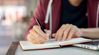 Masada dizüstü bilgisayarla otururken elinde tükenmez kalemle kırmızı bir deftere yazı yaz. Çalış ya da dışarıda çalış.