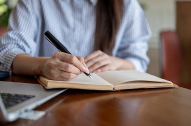 Kafedeki ahşap masada otururken, mavi çizgili gömlekli bir kadının dolma kalemle deftere yazdığını yakın plan. Dışarıda çalış ya da çalış.