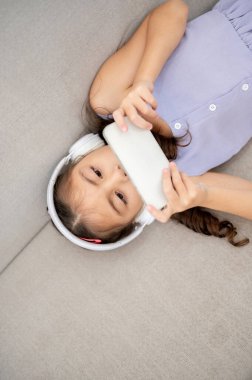 Asyalı bir çocuk ya da beyaz kulaklık takan küçük bir kız. Kumaş kanepede yatarken iki elinde de akıllı telefon tutuyor. Video izlemek ya da müzik dinlemek..