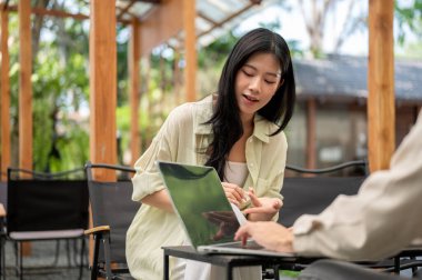 Asyalı kadın katlanır sandalyede otururken arkadaşıyla birlikte laptop ekranına bakmak için eğiliyor. Çalışmak ya da dışarıda okumak, Garden Cafe ya da Restoran.