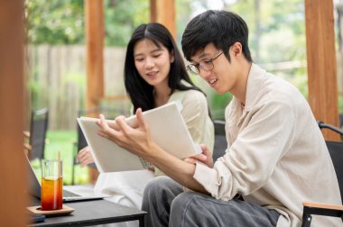 Gözlüklü Asyalı adam katlanan masadaki kız arkadaşına not defterini gösterirken gülümsüyor. Çalışmak ya da dışarıda okumak, Garden Cafe ya da Restoran.