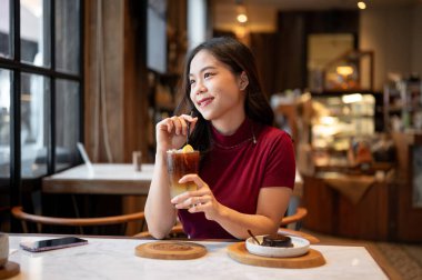Kamış tutan genç Asyalı kadın kafeteryada portakallı kahveyi yudumlarken ya da yan masada kek yerken. Yiyecek ve içecek, estetik kafe, tavsiye..
