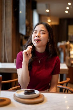 Asyalı genç bir kadın kaşık yalarken kafe masasında çikolatalı pasta yemekten zevk alıyor. Tatlı Tatlı Tatlı, Estetik Kafe, Dükkan Tavsiyesi.