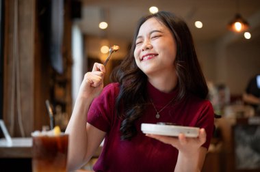 Genç Asyalı kadın kaşık tutarken gözlerini kapayıp gülümsüyor ve kafede yemek yiyor. Yiyecek ve içecek, estetik kafe, tavsiye..