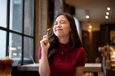 Genç Asyalı kadın, elinde kaşık tutarken gülümseyerek gözlerini kapatıyor ve kafede yemek yiyor. Tatlı Tatlı Tatlı, Estetik Kafe, Dükkan Tavsiyesi.