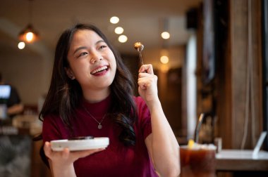 Genç Asyalı kadın kaşık tutarken ya da kaldırırken gülümseyerek gözlerini kapatıyor ve kafede yemek yiyor. Yiyecek ve içecek, estetik kafe, tavsiye..