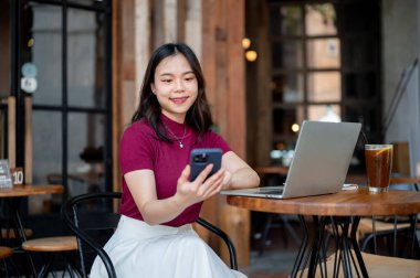 Asyalı bir kadın telefonu elinde tutuyor ve dizüstü bilgisayarını ve kahveyi kafedeki ahşap masada oturuyor. Çalışmak ya da Çalışmak, Serbest Çalışmak, Estetik Kafe.