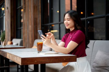 Kahvenin masasında kanepede otururken gülümseyen Asyalı kadın telefonu bir kenara koyup kahveyi işaret ediyor. Dışarısı soğuk, Estetik Kafe, Şehir Hayatı.