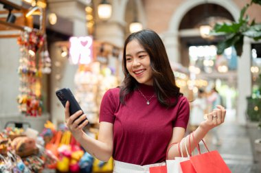 Alışveriş torbaları tutan Asyalı kadın bir markette ya da alışveriş merkezinde hediyelik eşya arabasının yanında telefona bakıyor. Ödeme günü tanıtımı, seyahat gezisi, turistik yerler..